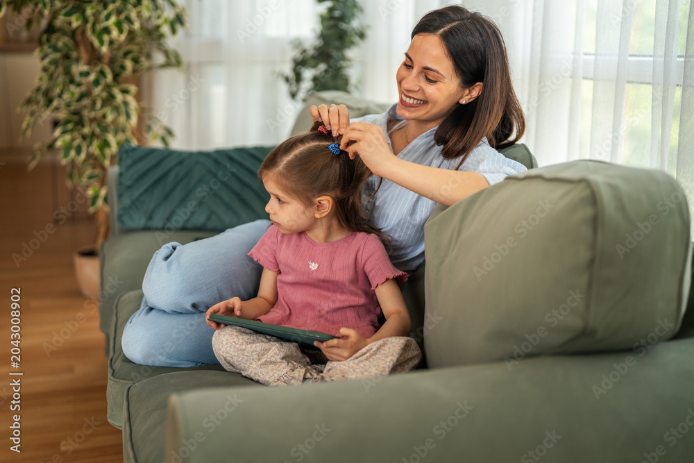 Fototapeta premium Mother styling daughter's hair while bonding on sofa