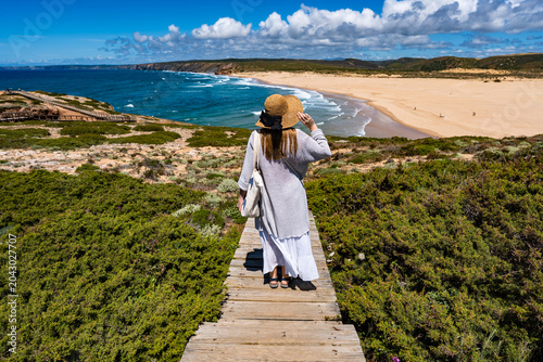 Beautiful middle-aged woman tourist walking down wooden stairs to sandy Bordeira beach and enjoying vacations in Algarve Portugal on summer day. Back view	