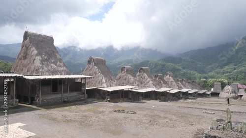 Panoramic view of Gurusina traditional village near Bajawa, Flores, Indonesia, featuring rows of iconic thatched houses beneath mist-covered mountains and lush tropical forest