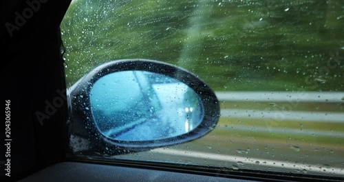 Interior point of view shot looking through a wet side window covered in rain droplets, focusing on a blue-tinted side view mirror as the vehicle drives past a blurred landscape of green hills