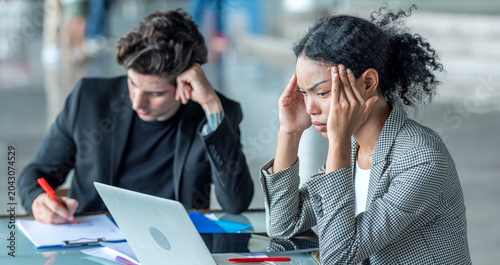 Stressed young businesswoman holding head working project about problem on laptop and documents during intense office discussion in business meeting with colleague workplace deadline pressure.