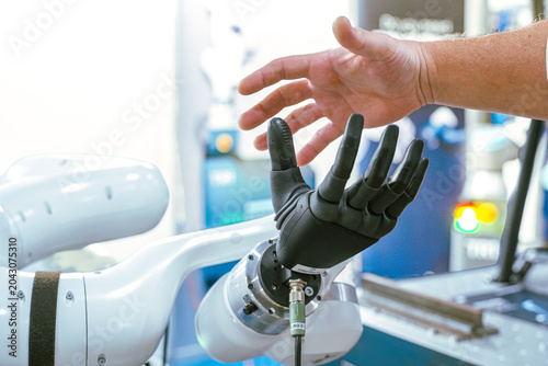 A human hand interacts with a multi-jointed robotic hand attachment on an automated mechanical arm during a demonstration of advanced industrial automation and cybernetic technology.