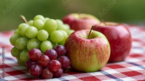 Fresh green and red grapes with ripe red apples arranged on a red and white checkered tablecloth, symbolizing healthy eating, seasonal harvest, and outdoor summer picnics