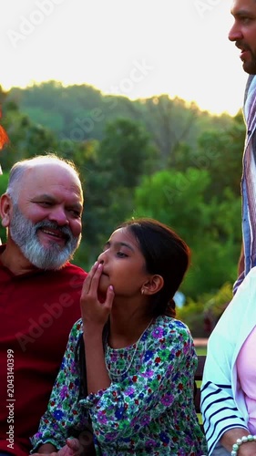 Indian family eating snacks while sitting on bench together outdoors in nature, smiling at camera, sharing joyful bonding moments, relaxed conversations, warm laughter, happy quality time outdoors