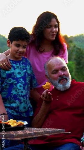 Indian family eating snacks while sitting on bench together outdoors in nature, smiling at camera, sharing joyful bonding moments, relaxed conversations, warm laughter, happy quality time outdoors