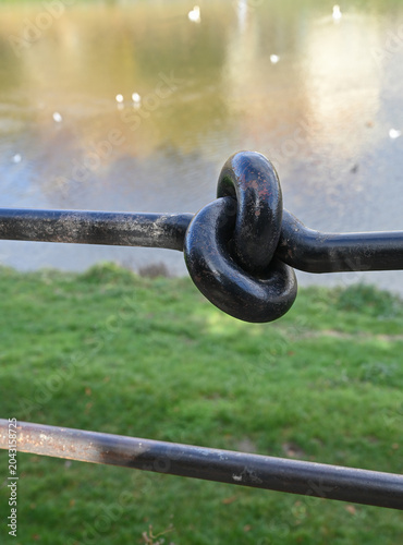 Close up of a twisted metal knot on a fence by a lake with grass in the background and small birds flying above