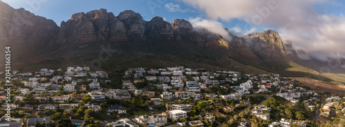 Bild auf Leinwand Aerial view of white modern homes on green slopes in Cape Town, South Africa, with the Twelve Apostles peaks under low clouds as late sun casts warm highlights and shadows