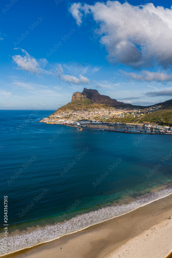 Obraz premium Aerial view of Hout Bay in Cape Town shows sandy beach, turquoise to deep blue water, curving harbor breakwater, rugged mountains, and late afternoon light under clouds.
