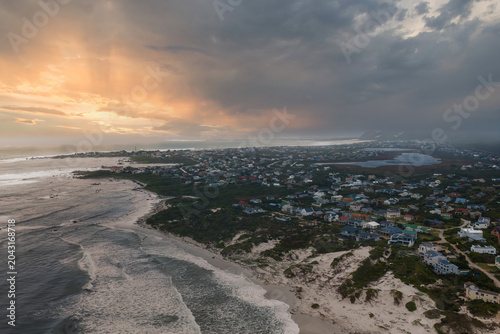 Aerial view of a coastal suburb near Cape Town as Atlantic surf meets a curving sandy beach and dunes, with rooftops by a lagoon and storm clouds glowing at sunset.