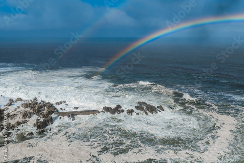 Aerial view of Cape of Good Hope near Cape Town, South Africa, as waves churn around dark rocks under a vivid rainbow and faint second arc after a daytime squall.