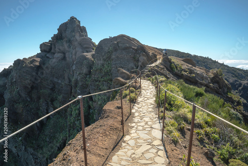 Fotografía A stone-paved walking trail with metal cable railings winds along a steep mountain ridge in Madeira, Portugal, surrounded by rugged volcanic rock and a clear blue sky