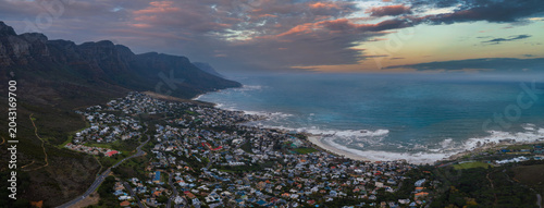 Aerial dusk view over Cape Town shows the Twelve Apostles on the left, surf lined bays, curving beaches, coastal neighborhoods, white sand, and gathering storm clouds.
