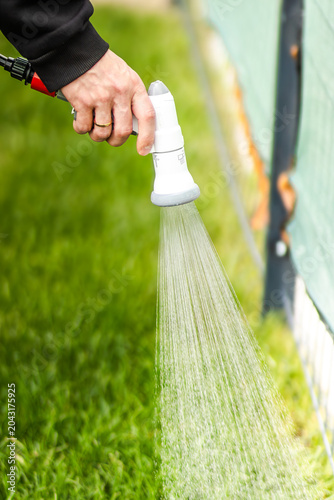 Hand holding a white garden hose nozzle spraying water onto green grass near a fence, showcasing a well-maintained outdoor area in a residential setting