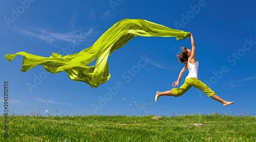 Vibrant young woman joyfully jumping in a lush green field on a bright sunny day holding a flowing lime green fabric with a clear blue sky background capturing energy freedom and happiness