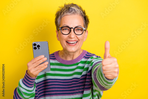 Fotografía Joyful mature woman with glasses holds smartphone and gives thumbs up against a