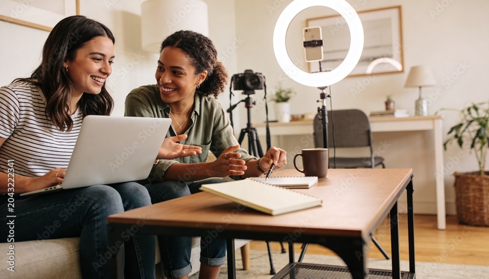 Fototapeta premium Reviewing two women in casual shirts, jeans leaning over laptop on sofa, ring light, copy space