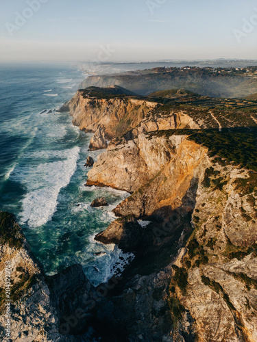 Aerial view of rugged coastal cliffs and powerful ocean waves crashing along a dramatic rocky shoreline at golden hour. Portugal. Cabo da Roca