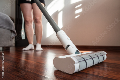 Photos Low angle shot of a white motorized floor brush of a cordless vacuum cleaner on a dark wooden floor, woman in white socks cleaning living room for perfect home hygiene and spotless lifestyle
