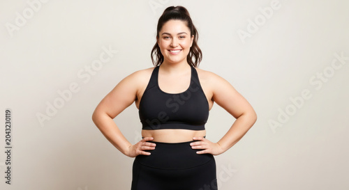 Plus-size woman in black sportswear poses confidently with hands on hips against a neutral background, showcasing fitness and body positivity