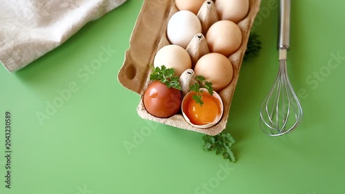 Fresh eggs in carton with whisk and herbs on green background