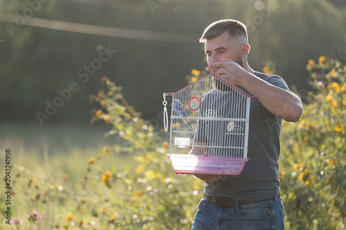Man gently holds a birdcage with a small bird inside, standing in a sunlit field.