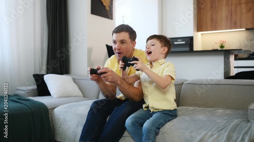 Father and young son enjoy playing video games on a sofa in a contemporary living room, with a kitchen area visible behind them