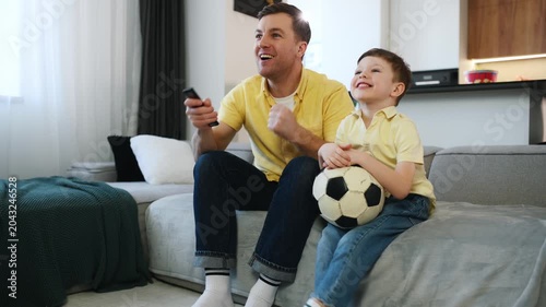 Father and young son relax on a couch in a living room, with the boy holding a soccer ball and the father holding a remote control