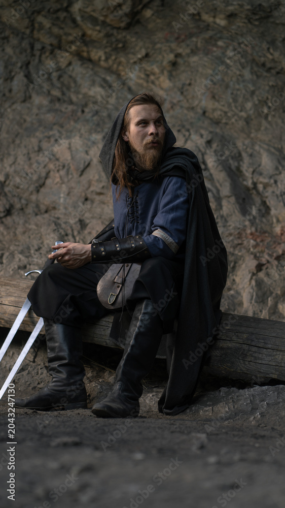 Fototapeta premium A weary warrior with a sword rests against a log, gazing thoughtfully into the distance.