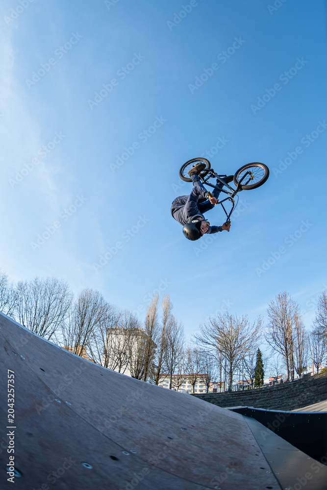 Naklejka premium Low angle shot of a young man doing an extreme flip stunt on a bicycle above a wooden ramp. The clear blue sky background emphasizes the high altitude and adrenaline of urban extreme sports.