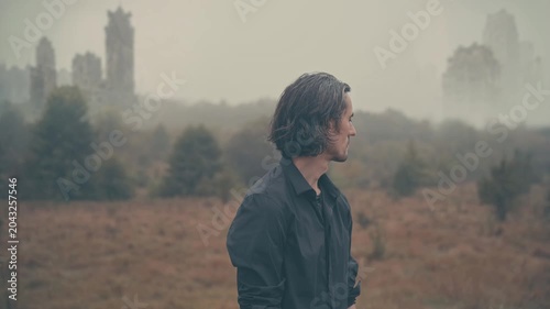 Lost man scanning foggy post-apocalyptic landscape with abandoned city, decayed ruins, dry grass, sparse bushes, sense of lost connection and collapsed civilization