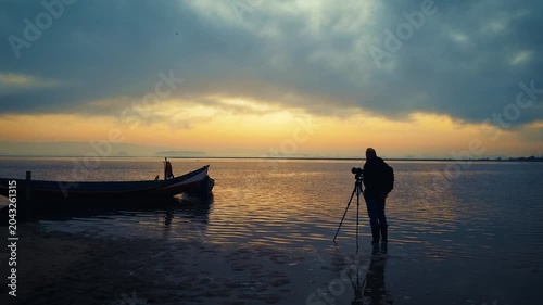 A photographer stands in shallow water with a tripod next to a traditional wooden fishing boat. The dramatic cloudy sky lights up with warm golden and blue tones during dawn.