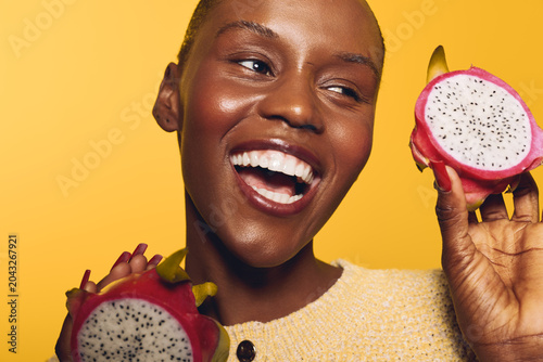Happy African woman with dark skin holding two halves of dragon fruit on yellow background. Close-up portrait with bright lighting and joyful expression. Concept of healthy lifestyle and freshness.