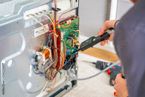 Technician testing electronic components and wiring inside disassembled electric oven. Appliance diagnostics and professional repair service concept