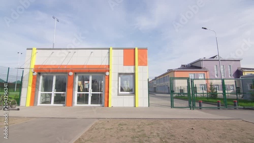 Entrance checkpoint building features colorful accents alongside glass double doors. Clean concrete pathway leading toward facility entrance