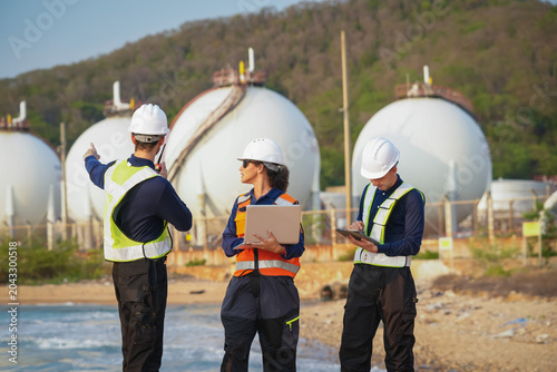 Safety inspection team analyzing gas storage facility coastal area business meeting industrial environment ground-level viewpoint