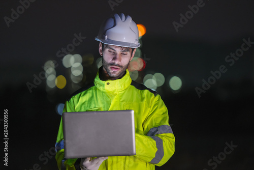 Nighttime industrial operations technician using laptop at oil refinery urban environment professional perspective safety focus