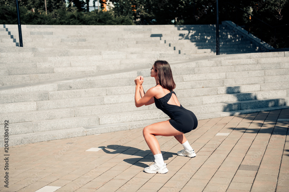 Naklejka premium Woman doing squat exercise outdoors on urban steps in athletic outfit and sneakers for fitness training and strength workout