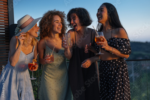 Young women friends laughing enjoying cocktails at sunset rooftop party