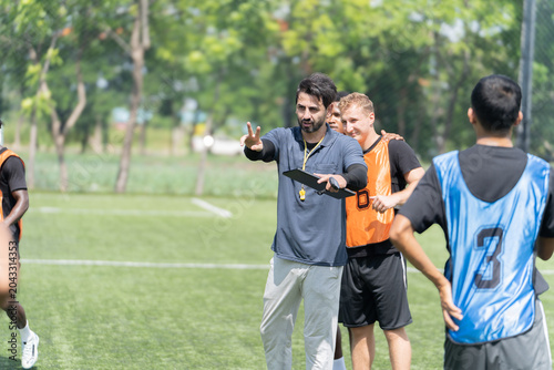 A referee with a whistle and clipboard gives instructions to multiethnic football players during an outdoor match on a sunny sports field. Multi ethnic group of soccer players on the soccer field