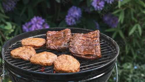 Foto Close‑up of a backyard charcoal grill with sizzling meat and burger patties cooking outdoors, surrounded by blooming purple flowers and greenery