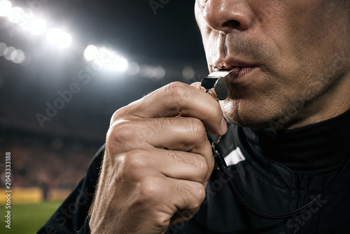 Close up of a football referee blowing a silver whistle during a match at night, professional soccer official making a call in a stadium under floodlights.