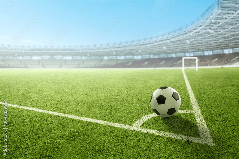 Fototapeta premium A black and white soccer ball on the corner kick line of a green field inside a large, empty stadium under a clear sky