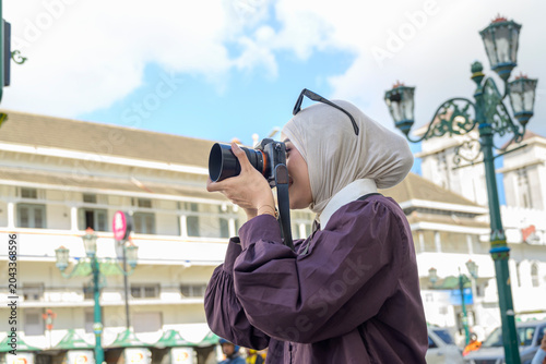 A modern Indonesian southeast asian muslim woman in a hijab takes photos with a digital camera while traveling in a historic city. Travel or vacation concept