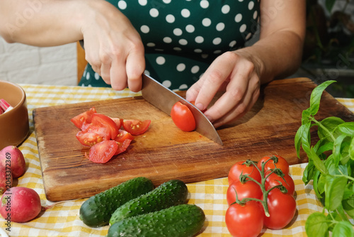 Cooking vegetable salad with tomatoes, cucumbers, garden radish and basil. Woman slicing fresh tomato in kitchen