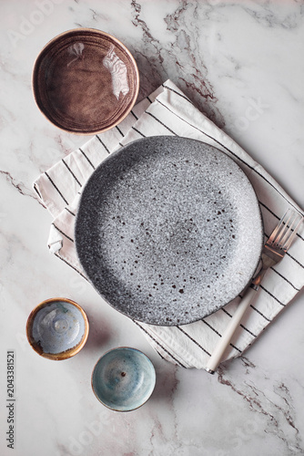 Top view flat lay of a minimalist table setting featuring a grey speckled ceramic plate, small bowls, a striped napkin, and a fork on a white marble surface.