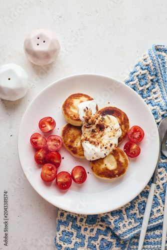 Savory syrniki (cheese pancakes) topped with sour cream, crispy fried onions, and fresh cherry tomatoes, served on a white plate with a blue knit cloth. Perfect for breakfast or brunch.