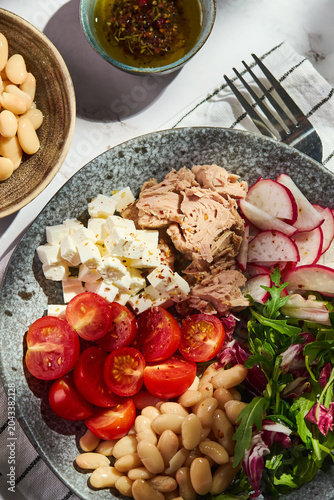 Top view of healthy tuna salad bowl with white beans, feta cheese, tomatoes, radish, arugula, and olive oil dressing on a marble surface. Top view