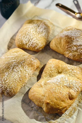 Close-up view of golden-brown homemade mini ciabatta rolls dusted with flour on parchment paper. Fresh artisan bakery bread with crispy crust texture.