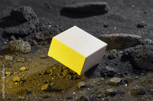White cardboard box sits amidst jagged charcoal rocks on a dark granular surface under harsh directional light that casts deep shadows and illuminates golden dust particles.
