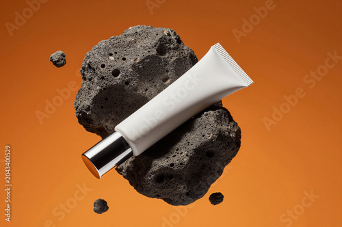 Cosmetic tube rests diagonally against a porous dark rock surface atop an orange backdrop. Sharp shadows fall across the textured stone while bright rim lighting accents the white packaging.
